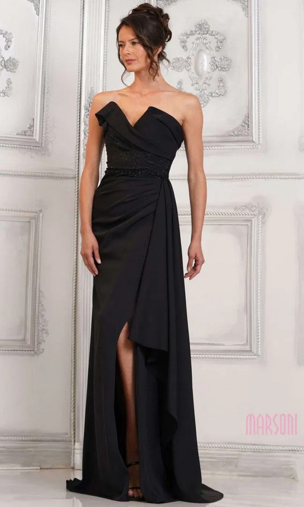 A woman stands indoors wearing the Marsoni MV1304—a black, strapless long formal dress with a ruffled neckline, floor-length silhouette, and an elegant beaded waist detail.